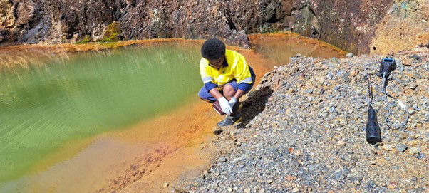 Miriama Toria Madigibuli kneeling next to extraction pit to collect water samples