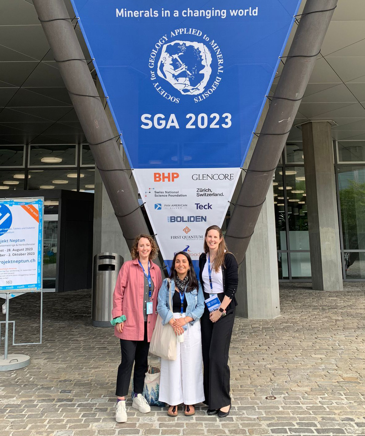 Rosie Blannin, Olivia Mejías, and Loren Nichols in front of  Society for Geology Applied to Mineral Deposits (SGA) banner